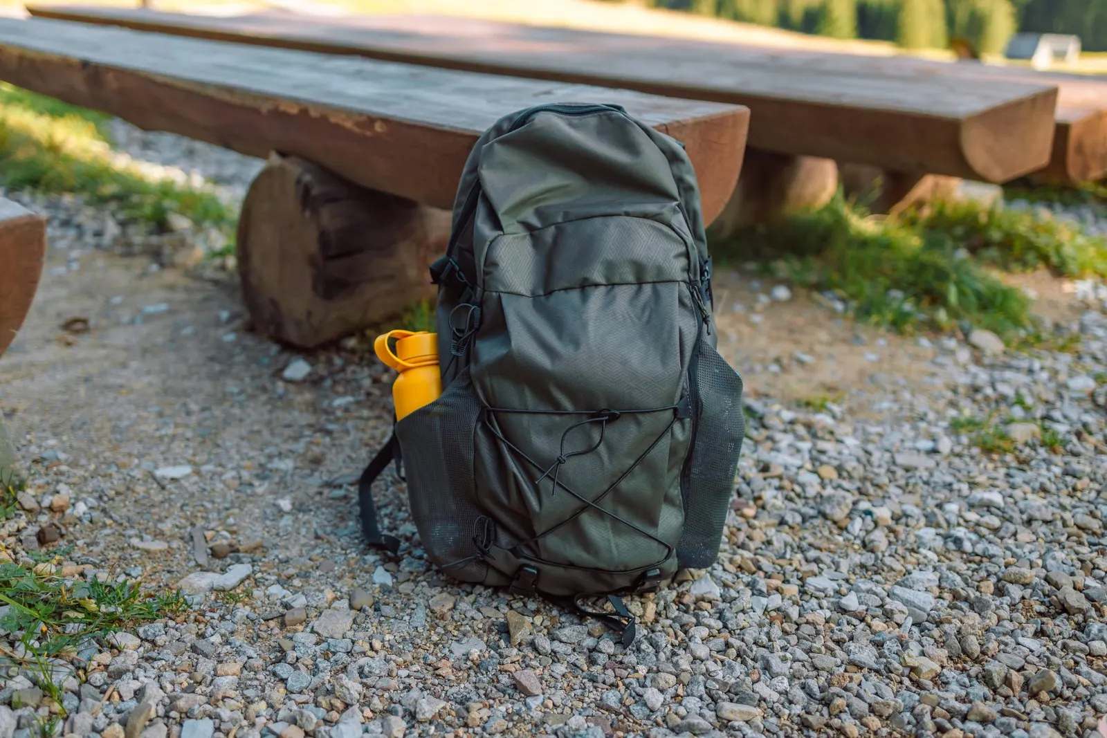 An olive backpack beside a wooden bench with a yellow bottle in a side pocket