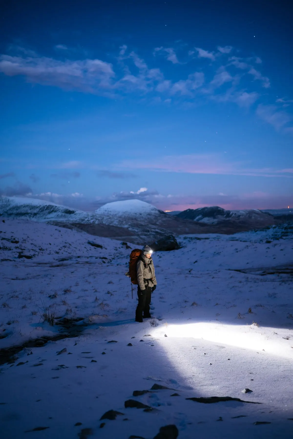 A hiker wearing a headlamp in a snowy landscape at twilight